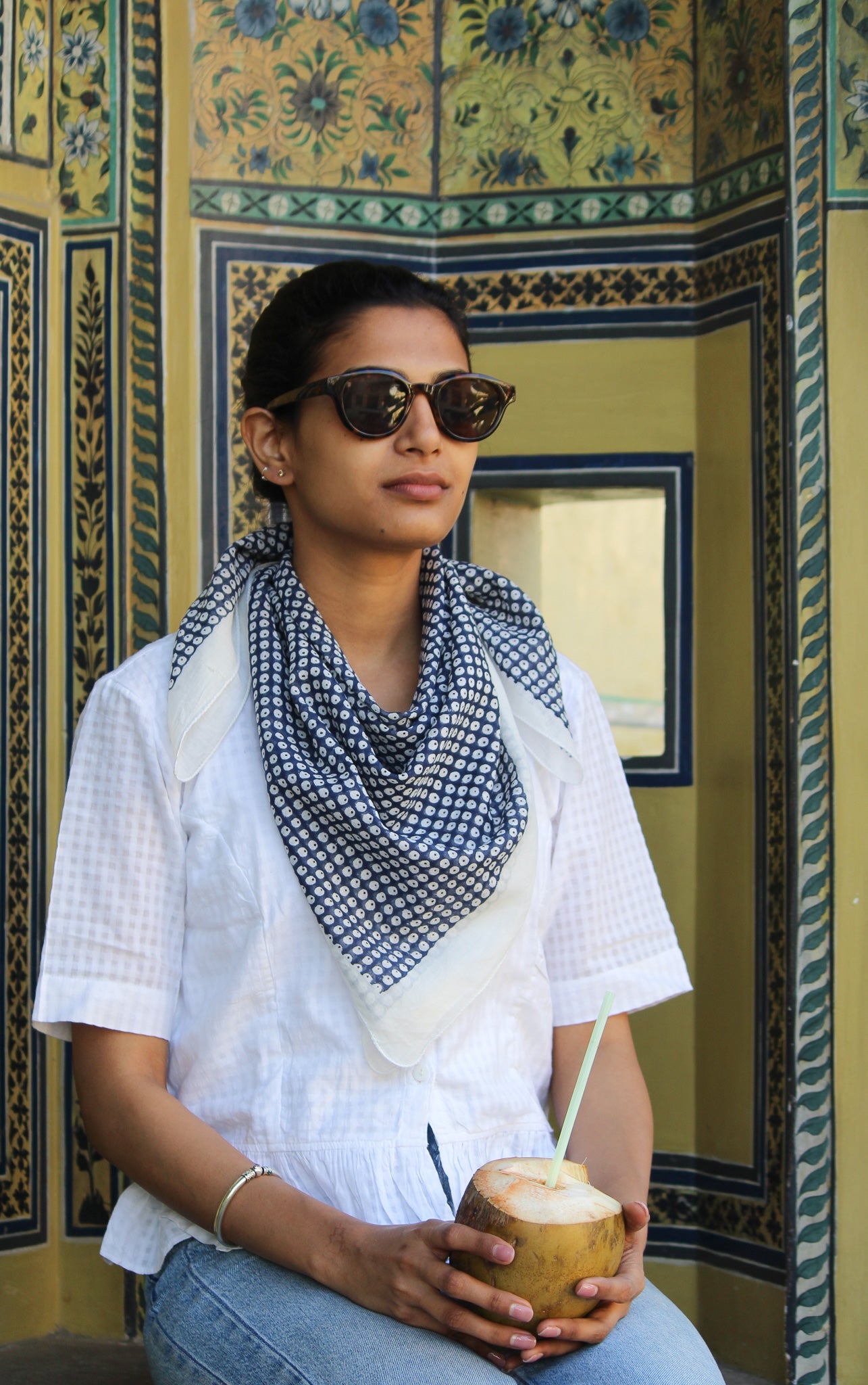 **Texte alternatif :** Jeune femme portant un foulard artisanal bleu et blanc en coton imprimé, assise devant un décor indien peint à la main, avec lunettes de soleil et noix de coco.